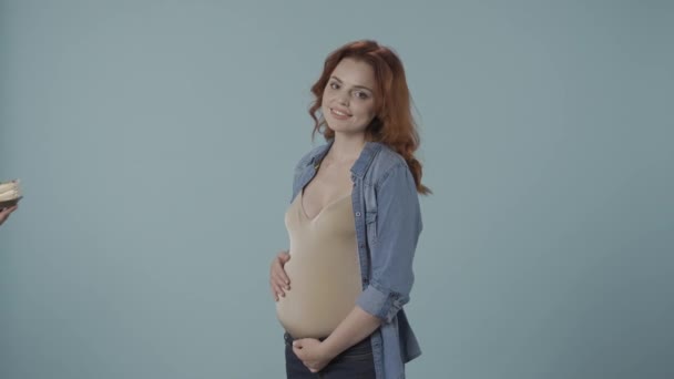 一个孕妇拿着一个盘子和一块甜蛋糕贪婪地看着它 一个带着一盘甜点的年轻孕妇的画像在工作室的蓝色背景 对怀孕的渴望 — 图库视频影像