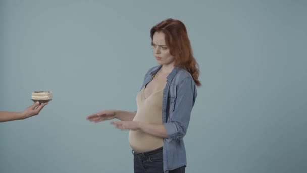 一个年轻的孕妇拒绝了一份带有蛋糕的盘子 一个女人在一个蓝色背景的工作室里做了一个停止的手势 怀孕期间的健康饮食和健康生活方式 — 图库视频影像