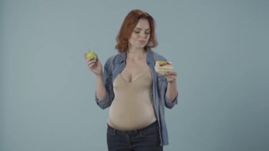 Hamile bir kadın sağlıklı ve sağlıksız yemek arasında seçim yapamaz. Bir kadın tatlı bir pasta ve yeşil bir elma arasında seçim yapar. Stüdyoda mavi arka planda bir kadın