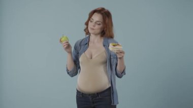 Hamile kadın sağlıklı ve sağlıksız yemek, pasta ve yeşil elma arasında seçim yapar. Kadın pasta lehine bir seçim yapar ve onu zevkle ısırır. Stüdyoda mavi arka planda bir kadın