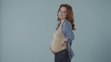 Kızıl saçlı hamile bir kadın şişmiş karnını gülümseyerek okşuyor, kızıyor, üzülüyor, alınıyor. Stüdyoda mavi arka planda bir kadın var. Hamilelik ve duygular, değişken ruh hali, hormonlar.