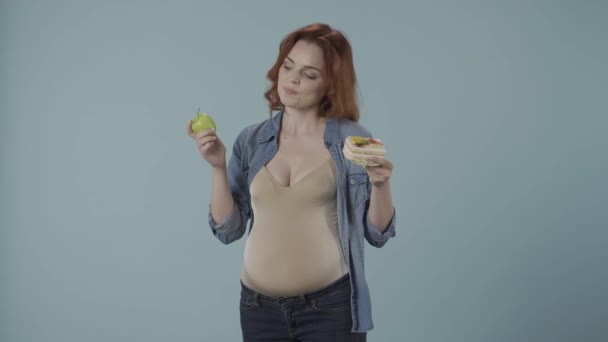 怀孕妇女在健康和不健康的食物 蛋糕和青苹果之间做出选择 女人做了一个有利于蛋糕的选择 高兴地咬着它 蓝色背景的工作室里的女人 — 图库视频影像