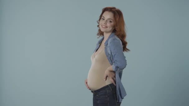 赤毛の妊婦が笑顔で彼女の膨らんだ腹を打ち 動揺し 怒った 青い背景のスタジオにいる女性 妊娠と感情 変化する気分 ホルモン — ストック動画