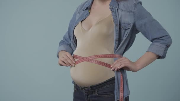 一个孕妇站在蓝色的背景下 用胶带测量她的腹部 怀孕妇女的腹部肿胀特写 怀孕期间检查体形和体形参数 — 图库视频影像