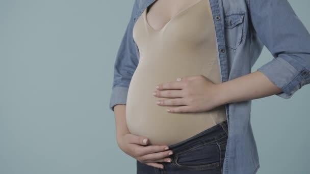 一个怀孕的女人用爱心和柔情抚摸着她臃肿的肚子 露出了一个大拇指 在一个蓝色背景的工作室里 一个怀孕妇女的腹部肿胀 怀孕的概念 — 图库视频影像