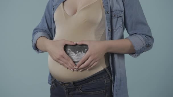 妊娠中の女性は 彼女の胃の近くに超音波スキャンを持っており 心臓の形をした手で折られています 青い背景にあるスタジオの妊婦の血まみれの腹 妊娠について — ストック動画