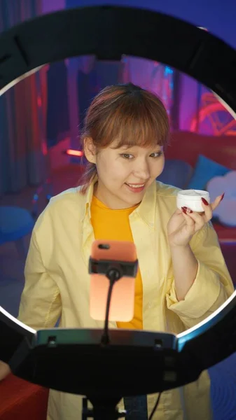 迷人的女性博主展示了一种新的面霜 在智能手机上拍摄了一篇评论 在彩灯前 女孩在家里录音室录制博客的肖像 垂直照片 — 图库照片