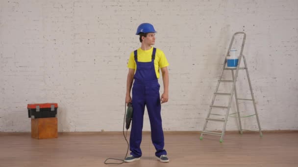 改装中の部屋に立っている笑顔の若い労働者の全長ビデオ ドリルを上げ 空気中の森林破壊者と彼の筋肉を示しています 会社広告について — ストック動画