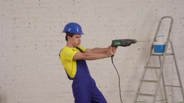 Tadilat altındaki odada elinde matkap, delgi, delgi ve matkap tutan genç bir inşaat işçisinin orta boy videosu. Tamir, imalatçı, şirket reklamı..