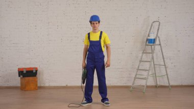 Elinde matkapla odada duran gülümseyen genç bir işçinin, elinde delgiyle kameraya bakarken tam boy çekimi. İnşaat, boya, imalatçı, şirket reklamı..