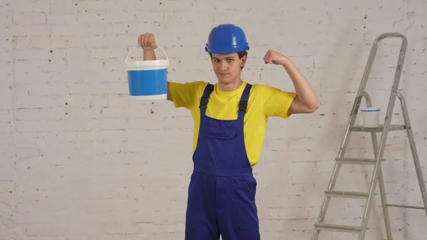 一个年轻的建筑工人站在正在翻新的房间里 拿着一桶油漆 露出他的肌肉 制造商 公司广告 — 图库照片