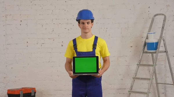 一个年轻的建筑工人站在房间里 拿着一个有广告区的笔记本电脑 工作空间被模仿着 面带微笑地看着 制造商 公司广告 — 图库照片
