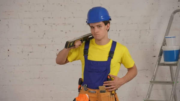 ツールベルトを身に着けている笑顔の若い建設作業員のミディアムショットは 彼の肩に建設水位を保持し 笑顔で自信を持ってカメラを見ています 会社広告について — ストック写真