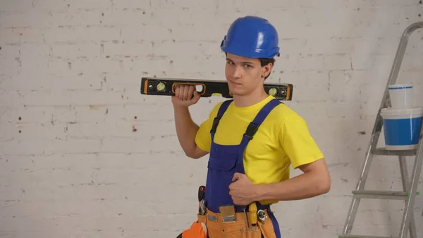 一名年轻的建筑工人戴着工具腰带 肩膀上抱着一个建筑水平面 微笑着 自信地看着相机 拍了一张中等的照片 公司广告 — 图库照片