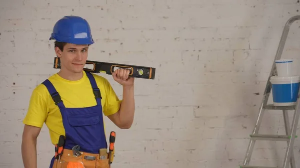 一名年轻的建筑工人戴着工具腰带 肩膀上抱着一个建筑水平面 微笑着 自信地看着相机 拍了一张中等的照片 公司广告 — 图库照片
