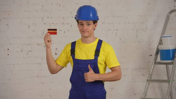 彼の手にクレジットカードで改装の下に部屋に立っている若い建設労働者の中型ショット 親指を与える 製品を推奨 プロダクト 会社広告 — ストック写真