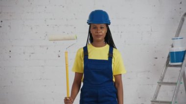 Koyu tenli genç bir kadın işçinin elinde silindirle odada durup, yeni boyanmış duvarlara bakarken ve baş parmağını kaldırırken orta boy videosu. İnşaat, boya, imalatçı, şirket reklamı.