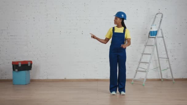 一个身穿制服 头戴硬礼帽的深色皮肤女建筑工人站在房间里指着边框的全长视频 你的广告位置 — 图库视频影像