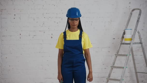 改装中の部屋に立っているダークスキンの若い女性建設労働者の中型ビデオは 壁とローラーのためのペンキでいっぱいのバケツを示しています ペイント 企業広告 — ストック動画