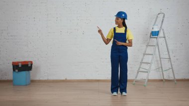 Koyu tenli, üniformalı ve kasketli genç bir kadın inşaat işçisinin çerçevenin yan tarafını gösteren tam boy fotoğrafı. Reklamınız için yer açın. İnşaat, şirket.