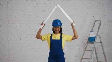Koyu tenli genç bir kadın inşaat işçisinin başının üstünde iki adet katlanabilir plan büyütürken, bir çatı taklidi yaparken orta boy fotoğrafı. İnşaat, imalatçı, şirket reklamı. Güvenlik, güvenilirlik.