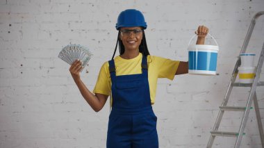 Koyu tenli, genç bir inşaat işçisinin bir kova boya ve bir sürü banknot göstererek odanın içinde dikilirken orta ölçekli bir fotoğrafı. Boya, şirket reklamı..