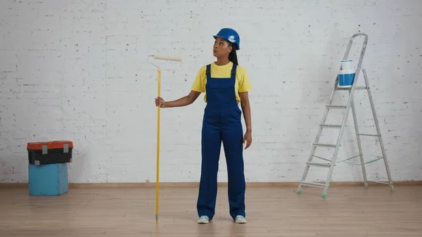 一个皮肤黝黑的年轻女建筑工人拿着一个滚筒站在房间里 看着新刷好的墙壁 制造商广告 — 图库照片
