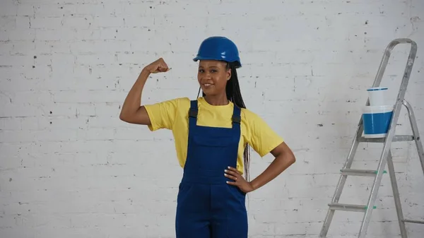 彼女の筋肉を示す改装の下で部屋に立っているユニフォームと硬い立場の笑顔の暗い肌の若い女性の建設労働者の中型ショット メーカー 企業広告 — ストック写真