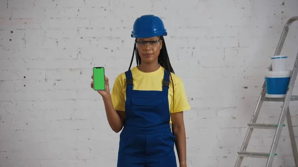 リノベーションの下に部屋に立っているダークスキンの若い女性建設労働者の中型ショットは 広告エリアを持つスマートフォンを保持し ワークスペースがモックアップします モバイルアプリ — ストック写真