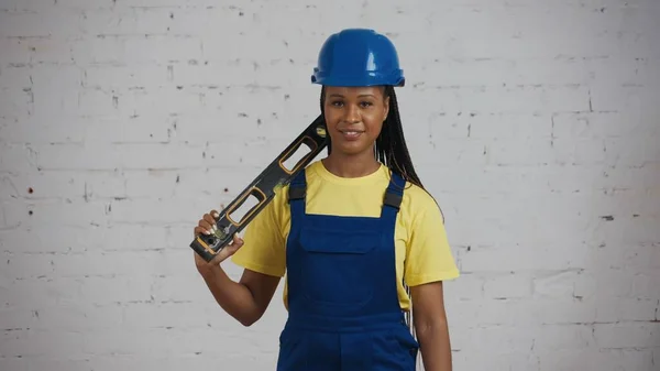 一名深色皮肤的年轻女建筑工人站在正在翻新的房间里 肩膀上顶着一个建筑水平面 面带微笑地拍了照 — 图库照片