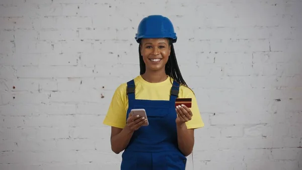 一名皮肤黝黑的年轻女建筑工人手持信用卡和电话 站在正在翻新的房间里 拍了一张中景照片 公司广告 — 图库照片