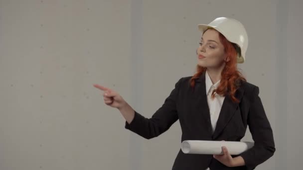 女性建築家は何かを指摘する グレーの背景にスタジオで彼女の手の青写真を持つヘルメットの女性の肖像画 プロモーション — ストック動画