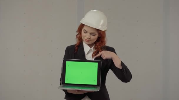 女性アーキテクトが前に向かってラップトップスクリーンを保持し 緑色の画面を指しています スタジオで灰色の背景に彼女の手のハード帽子とラップトップでビジネス女性の肖像画 — ストック動画