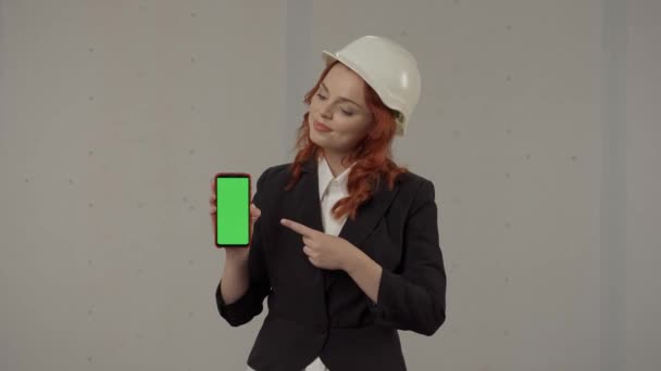 女性の建築家がスマートフォン画面を前面に持ち 緑色の画面を指差しています スタジオの灰色の背景にヘルメットでビジネス女性の肖像画 広告エリア — ストック動画