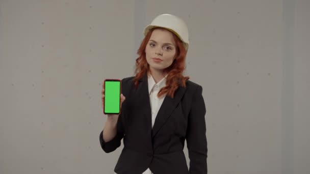 女性アーキテクトが顔を向けてスマートフォン画面を保持し 緑色の画面を指差し 親指を示しています ヘルメットのビジネス女性の肖像画とグレーの彼女の手のスマートフォン — ストック動画