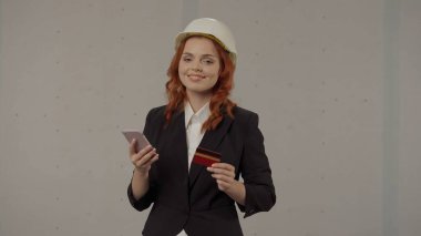 Bir kadın mimar, akıllı telefon ve kredi kartıyla online bir mağazadan satın almak için ödeme yapıyor. Stüdyoda, gri arka planda, koruyucu kasklı bir iş kadını portresi.