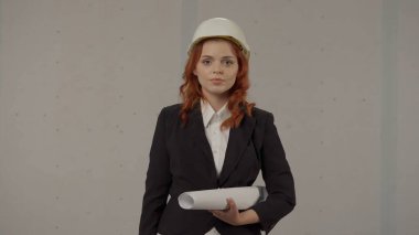 Elinde planları olan genç bir mimarın portresi. Yakın plan. Koruyucu kasklı bir kadın mimar ve gri arka planda stüdyoda sıkı bir takım elbise. Mimari proje