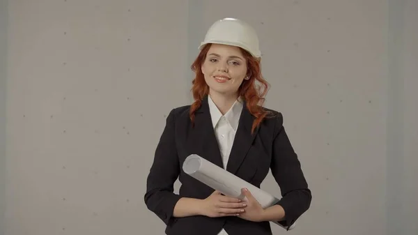 一个手里拿着设计图的年轻女建筑师的画像 女建筑师戴着防护头盔 穿着紧身衣 穿着灰色背景的工作室 建筑项目 — 图库照片