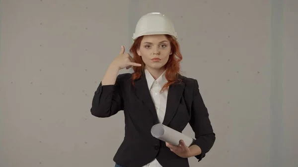 一位女建筑师在她的头部附近做了一个电话手势 一个头戴硬礼帽 手里拿着蓝图 背景灰暗的女人的画像 — 图库照片