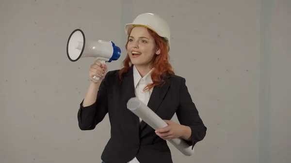一个戴着防护头盔 手里拿着画的女人对着牛角说话 一位女建筑师在灰色背景的工作室里用扩音器校正作品 — 图库照片