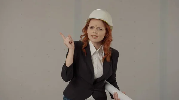 Architect woman gets angry, speaks emotionally, shakes her index finger. Portrait of a woman in a protective helmet and with blueprints in the studio on a gray background
