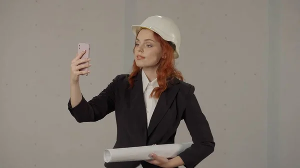 女建筑师通过视频通话 用智能手机自拍 一个戴着头盔 在工作室里有灰色背景蓝图的女人 — 图库照片