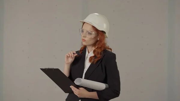 一个戴着头盔 腋下夹着设计图的女人在工作室里 背景是灰色的 一位戴防护眼镜的女建筑师想得很周到 — 图库照片