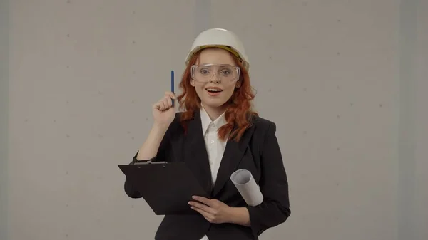 A woman in a helmet and with blueprints under her arm in the studio on a gray background. A woman architect in protective glasses happily exclaims there is an idea