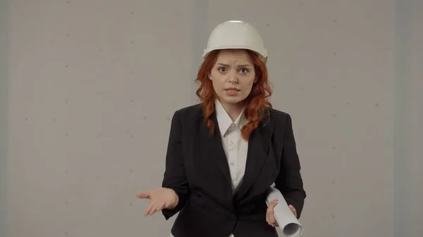 Angered woman speaks emotionally, gesticulating with her hands. Portrait of a woman in a protective helmet and with blueprints in the studio on a gray background