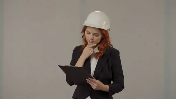 A woman in a helmet and with blueprints under her arm in the studio on a gray background. A woman architect is thinking hard while developing a new project