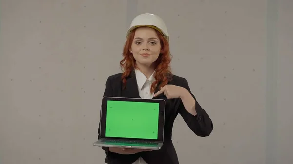一位女建筑师拿着一个面向前方的笔记本电脑屏幕 指着它的绿色屏幕 工作室里一个头戴硬礼帽 手里拿着笔记本电脑 背景灰暗的女商人的画像 — 图库照片