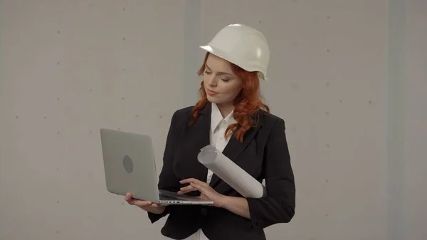 A woman architect works on a laptop, studies plans, prints reports. Business woman in a protective helmet, with drawings under her arm and a laptop on a gray background in the studio