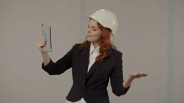 ビデオ通話で話す建築家の女性は タブレットを使用してセルフィーを取ります グレーの背景にスタジオで保護ヘルメットと厳格なスーツでビジネス女性 — ストック写真