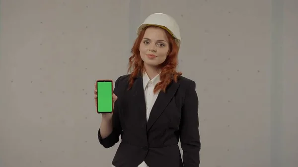 女建筑师展示绿色屏幕智能手机 工作室里一个戴着灰色背景头盔的女商人的画像 广告区 模拟工作区 — 图库照片